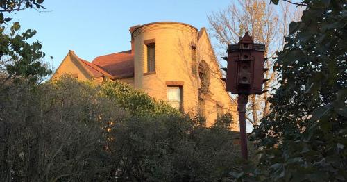 A historic brick house partially obscured by trees, with a decorative birdhouse in the foreground.