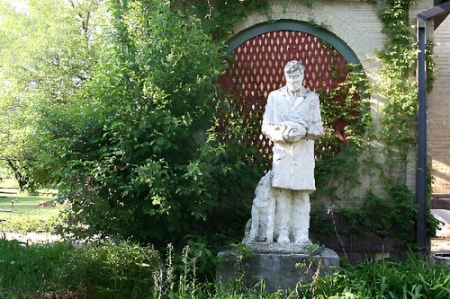 A white statue of a man holding a book, surrounded by greenery and a red lattice backdrop.