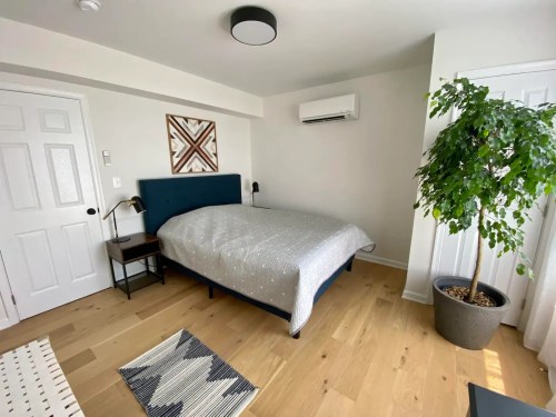 A cozy bedroom featuring a bed, nightstand, plant, and artwork on the wall, with light wood flooring.