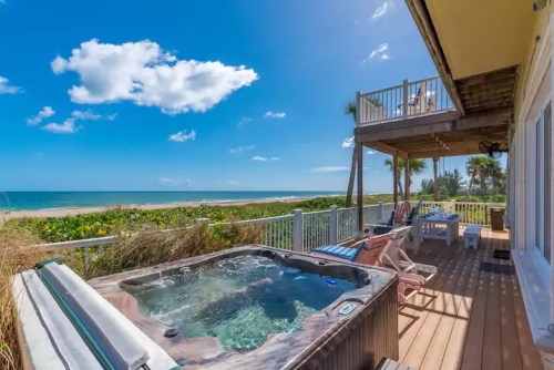 A beachfront deck with a hot tub, lounge chairs, and a view of the ocean under a clear blue sky.