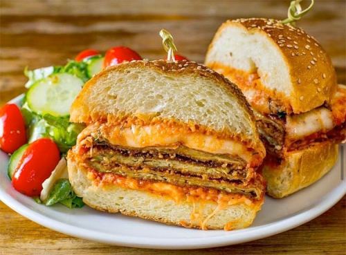 A close-up of a layered sandwich with chicken and sauce, served with a side salad of cherry tomatoes and cucumbers.