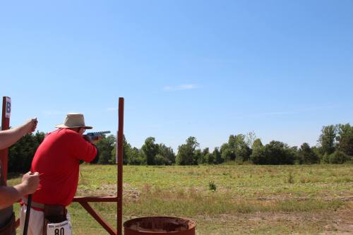 A person in a hat aims a rifle at a target in an open field under a clear blue sky.