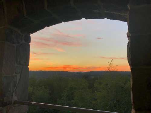 View of a colorful sunset through a stone arch, framed by trees and hills in the distance.