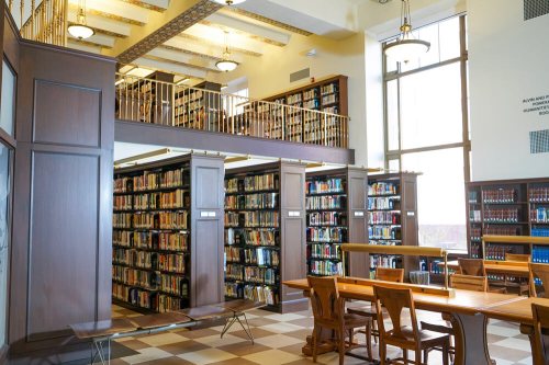 A spacious library interior with wooden shelves filled with books, tables for study, and large windows letting in natural light.