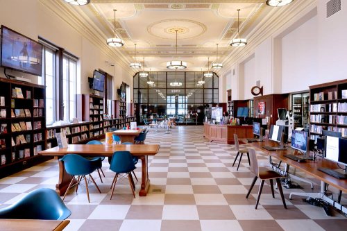 A spacious library interior with bookshelves, tables, computers, and large windows, featuring a checkered floor.