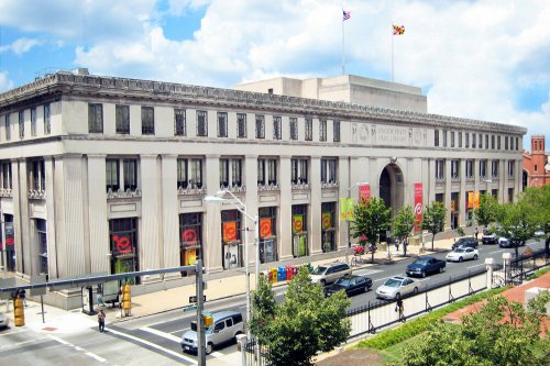 A large, historic building with columns, flags, and colorful banners, surrounded by trees and cars on a sunny day.