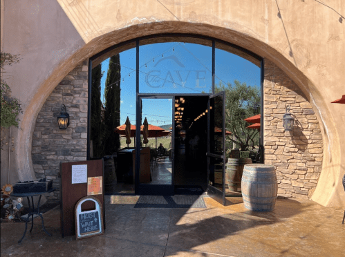 Entrance to "The Cave" restaurant, featuring large glass doors, outdoor seating, and decorative barrels.