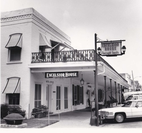Black and white photo of the Excelsior House hotel entrance with a sign, awnings, and parked cars.