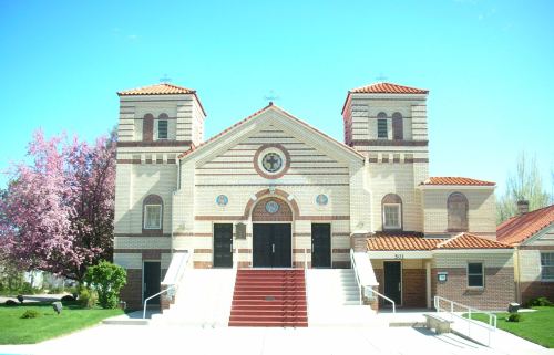 A brick church with a red-tiled roof, flanked by trees and a clear blue sky. Steps lead to the main entrance.