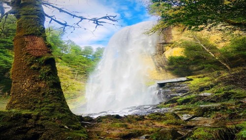A majestic waterfall cascades down rocky cliffs, surrounded by lush greenery and a clear blue sky.