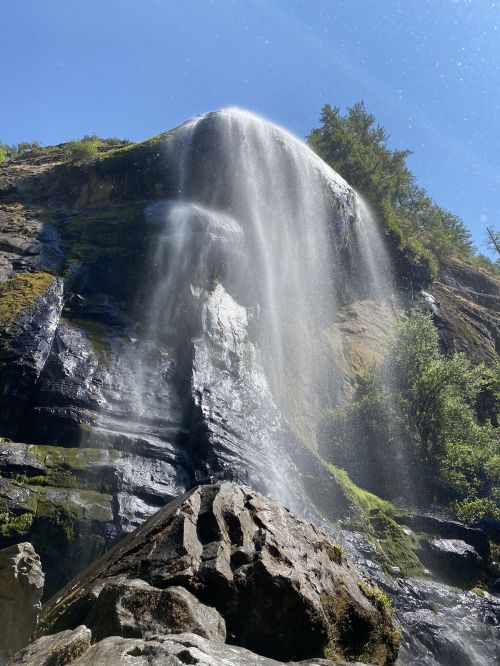 A waterfall cascading down a rocky cliff, surrounded by lush greenery and mist in the sunlight.