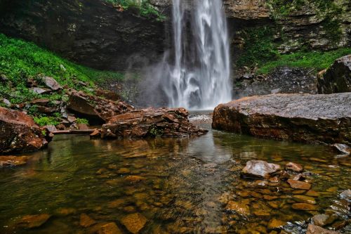 A serene waterfall cascades over rocks into a clear pool, surrounded by lush greenery and rugged terrain.