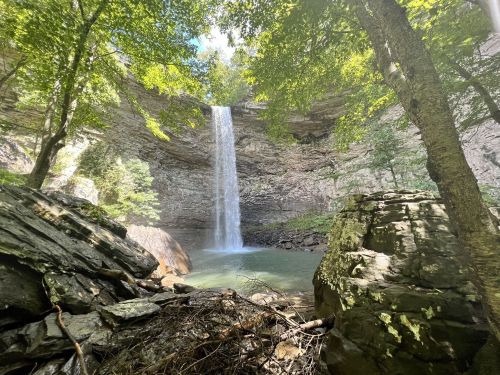 A serene waterfall cascades into a clear pool, surrounded by lush greenery and rocky terrain.