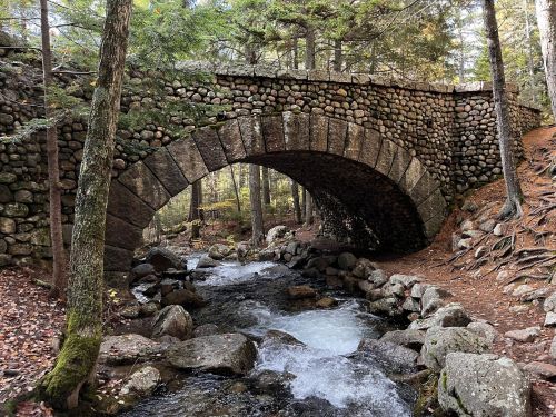 A stone arch bridge spans a flowing stream, surrounded by trees and rocky terrain in a serene forest setting.