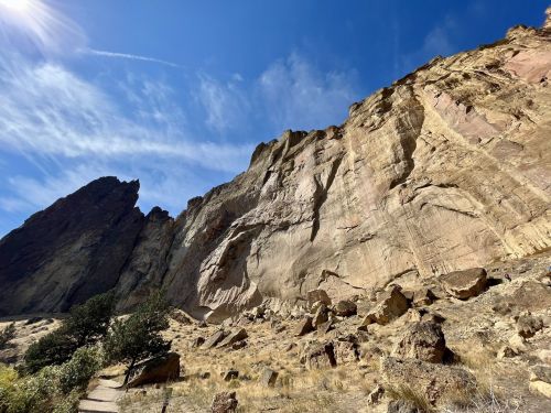 A rocky cliff rises against a blue sky, with sparse vegetation and a clear path in the foreground.