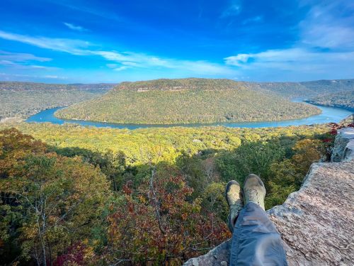 A person’s feet resting on a rocky ledge, overlooking a lush valley and winding river under a blue sky.