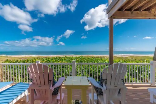 A beachfront view from a porch with colorful chairs, overlooking the ocean and a clear blue sky with clouds.