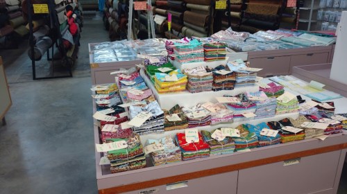 A display of colorful fabric bundles stacked on a table in a fabric store, with various patterns and labels visible.
