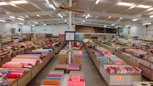 A spacious fabric store with rows of colorful fabric rolls and bolts organized on shelves, under bright lighting.