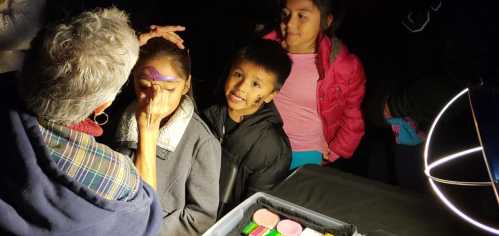 A child is getting their face painted while two others watch, with colorful face paint supplies visible nearby.