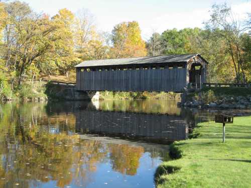 A wooden covered bridge spans a calm river, surrounded by colorful autumn trees and a grassy bank.