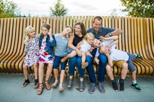 A family of seven poses playfully on a wooden bench, with kids making funny faces and adults smiling.