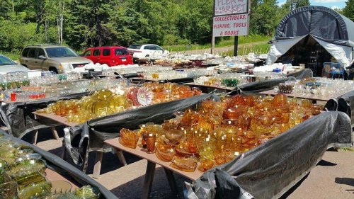 A bustling flea market with tables filled with colorful glassware and collectibles, surrounded by parked cars and greenery.