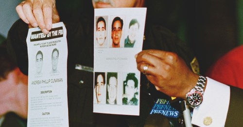Two men hold up wanted posters with photos and descriptions, speaking at a press conference.