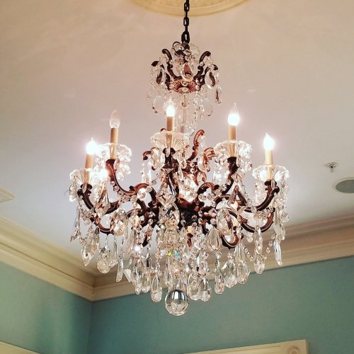 A decorative chandelier with crystal accents and six lit candles, hanging from a ceiling in a softly colored room.
