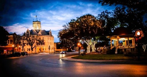 A charming town square at dusk, featuring a lit gazebo and historic building, surrounded by trees adorned with lights.
