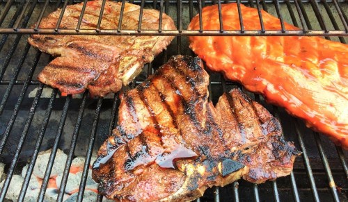 Grilled meats on a barbecue grill, including steaks and a rack of ribs, with visible grill marks and a smoky appearance.