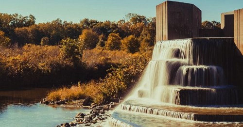A serene waterfall cascades over a stone structure, surrounded by autumn foliage and a calm river.