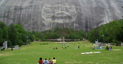 Visitors relax on a grassy area in front of a large stone carving on a mountain, surrounded by trees.