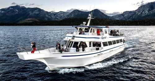 A large white boat with passengers cruising on a lake, surrounded by mountains under a cloudy sky.