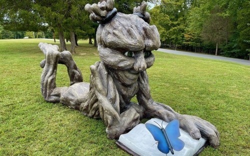A whimsical sculpture of a tree-like figure reading a book with a blue butterfly perched on it, set in a grassy park.