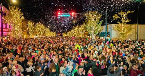 A large crowd gathers at night, surrounded by festive lights and falling snow, celebrating a winter event.