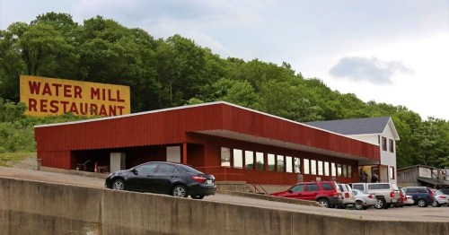 A modern restaurant building with large windows, set against a green hillside, featuring a prominent sign that reads "Water Mill Restaurant."