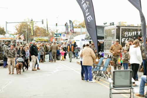 A busy outdoor event with people in camouflage, vendors, and dogs, set on a street with flags and tents.