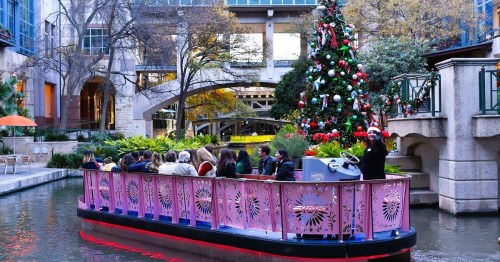 A festive boat with passengers glides along a decorated river, surrounded by greenery and a Christmas tree.