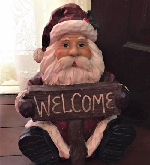 A decorative Santa figure holding a wooden "WELCOME" sign, sitting on a wooden floor near a door.