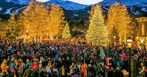 A bustling crowd gathers around a beautifully lit Christmas tree in a snowy mountain town during the holiday season.