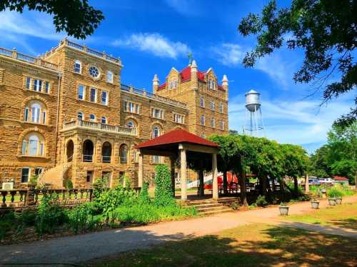 Historic stone building with ornate architecture, surrounded by greenery and a gazebo, under a bright blue sky.