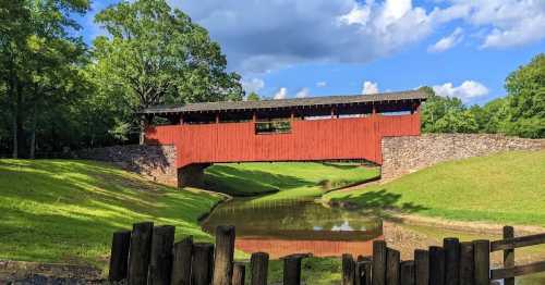 A red covered bridge spans a calm stream, surrounded by lush green grass and trees under a blue sky with clouds.