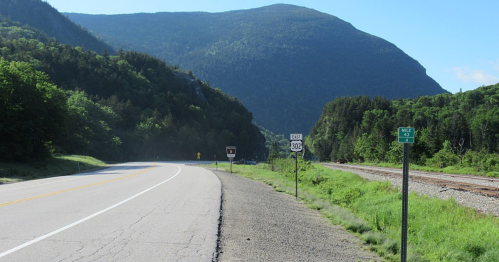 A scenic view of a winding road alongside a railway, with mountains in the background and mile marker signs visible.