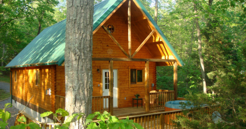 A cozy wooden cabin with a green roof, surrounded by trees and featuring a porch and hot tub.