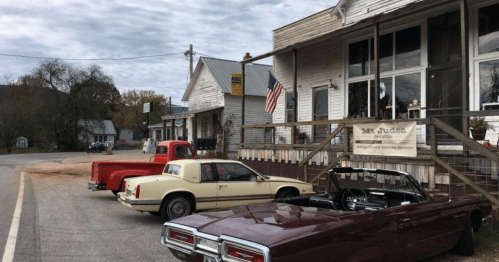A quaint roadside scene featuring vintage cars parked outside a rustic building with an American flag.