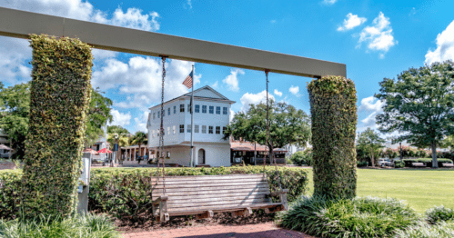 A wooden swing hangs from a frame, framed by greenery, with a historic building and blue sky in the background.