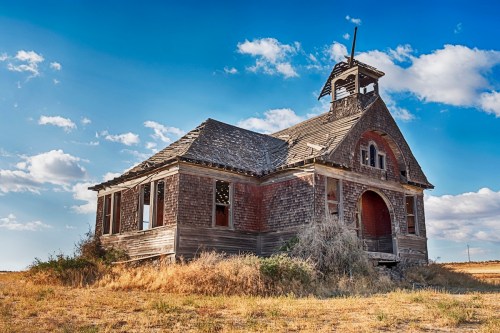 An abandoned, weathered schoolhouse with a bell tower, surrounded by dry grass and under a blue sky with clouds.