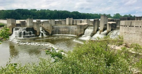 A concrete dam with cascading waterfalls surrounded by greenery and trees under a cloudy sky.
