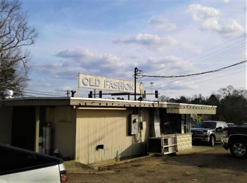 A small, vintage building with a sign reading "OLD FASHION" under a cloudy sky, surrounded by parked vehicles.